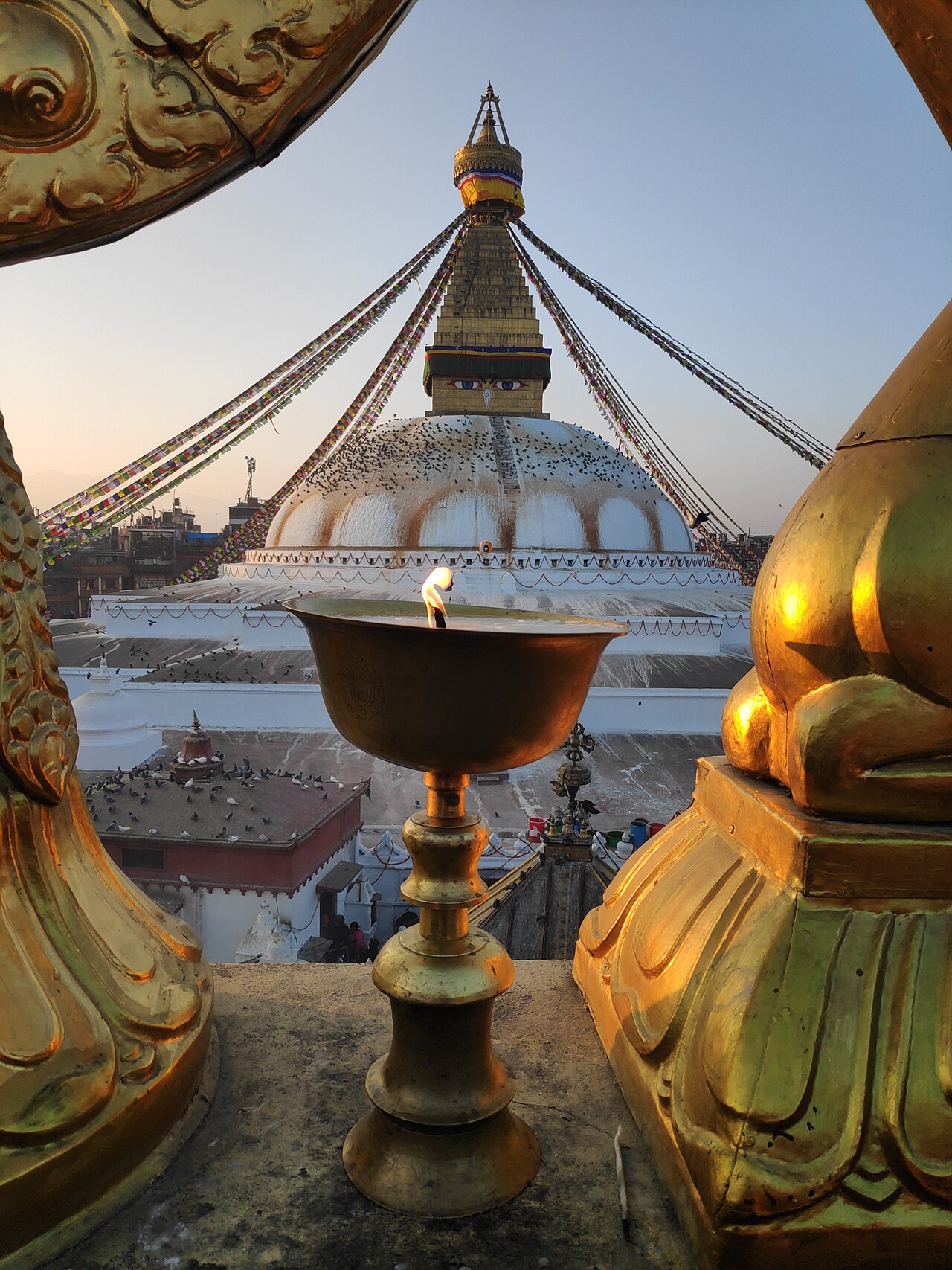 A butter lamp burning on a monastery terrace at dusk, the dome and spire of Boudhanath rising behind it