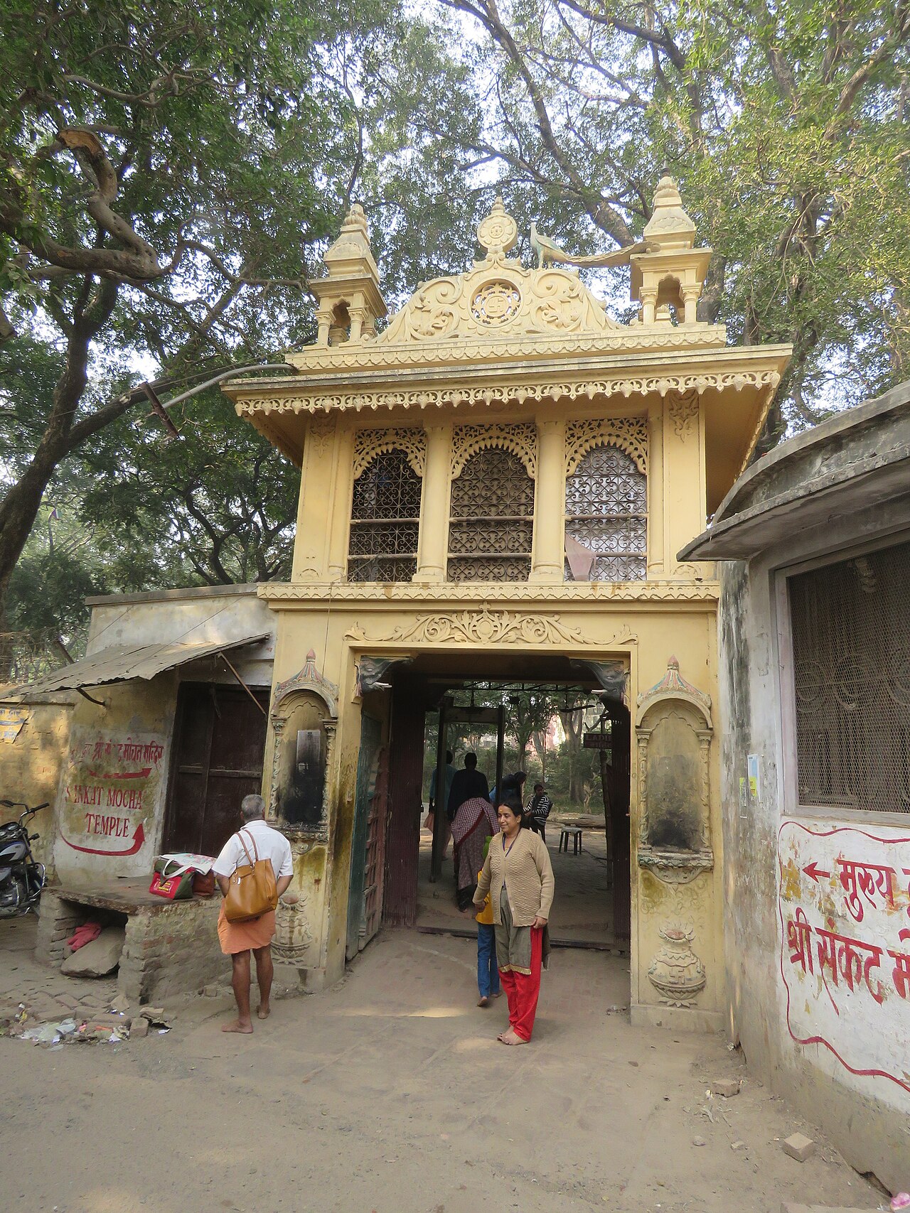 The Temple's ornamented yellow entrance gateway, with its Sankat Mochan Temple signboard and an offerings counter beside the steps