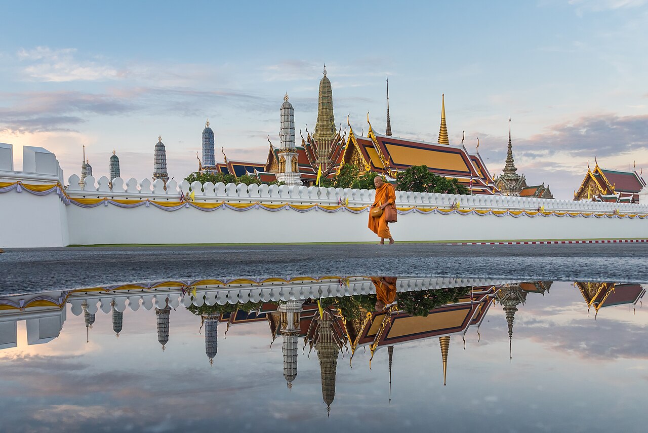 A monk in orange robes walks past the white crenellated wall of Wat Phra Kaew at dawn, the spires reflected in rainwater