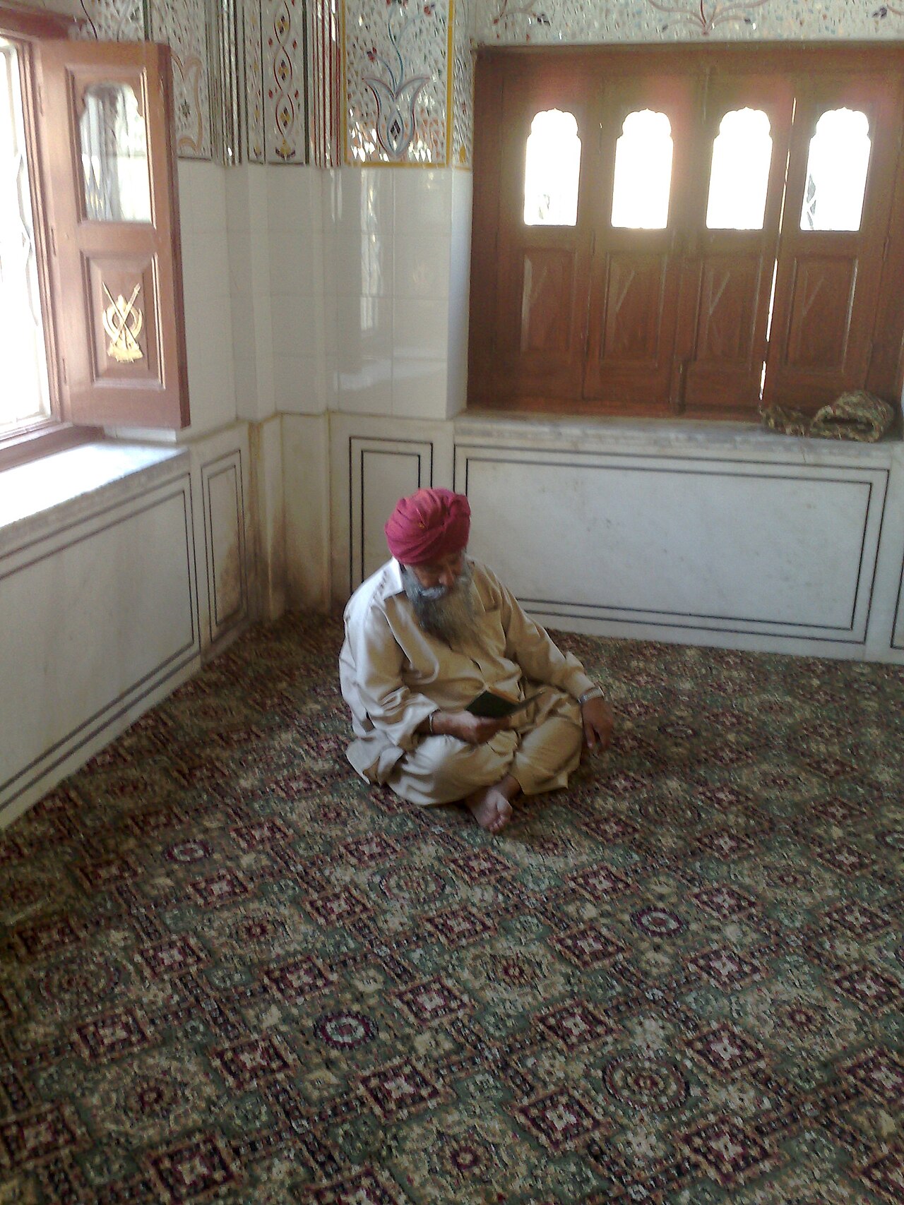 An elder reads from a Gutka Sahib in a marble chamber of Gurdwara Panja Sahib, a carved Khanda set into the door beside him