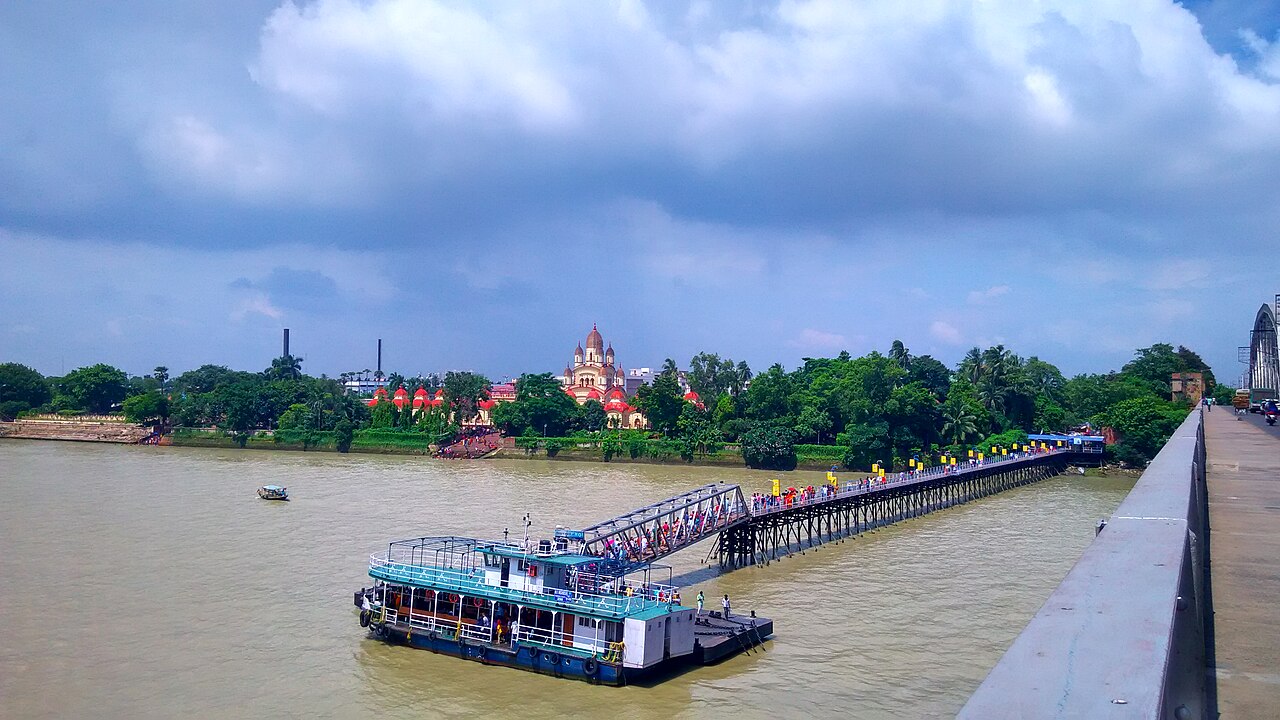 Pilgrims crossing the jetty walkway to the ferry at Dakshineswar, with the temple and the twelve Shiva shrines behind