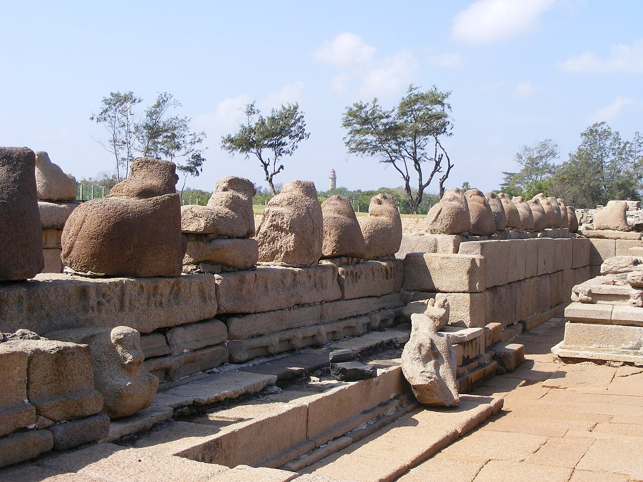 Rows of weathered Nandi bulls seated along the boundary wall of the Shore Temple, with the Mahabalipuram lighthouse in the distance
