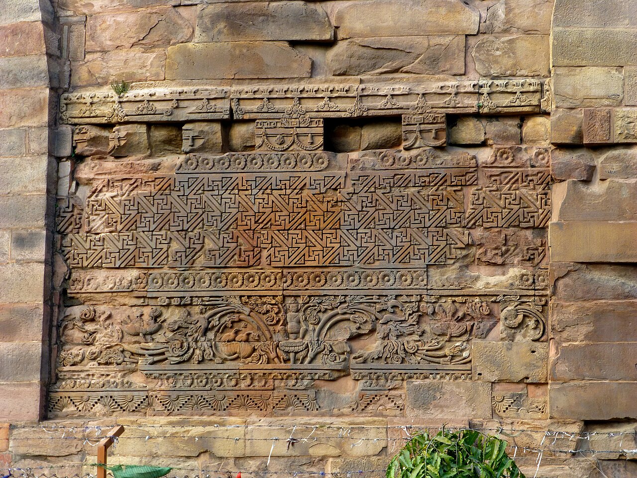 Close-up of Gupta-period stone carving on the Dhamek Stupa's base, showing geometric key-pattern bands, lotus rosettes and carved figures
