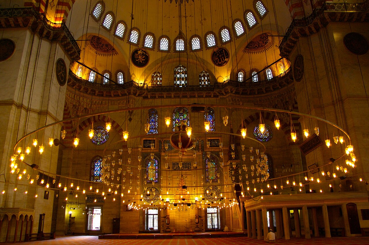 The interior of the Süleymaniye Mosque, the low iron ring of oil lamps hanging beneath the great dome and its ring of windows, with the tiled qibla wall beyond