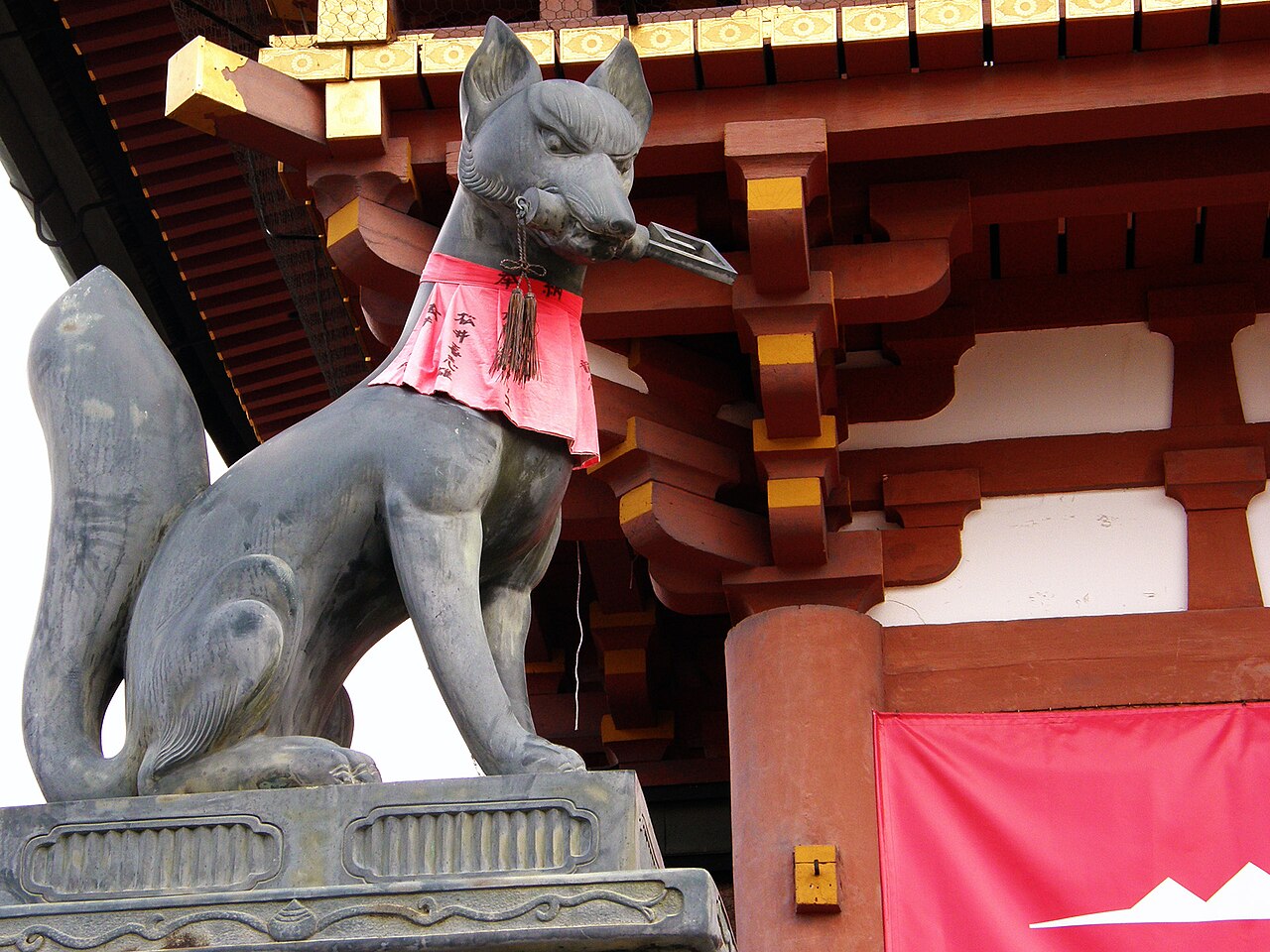 Stone fox messenger with a granary key in its teeth and a red votive bib, at the gate of Fushimi Inari Taisha