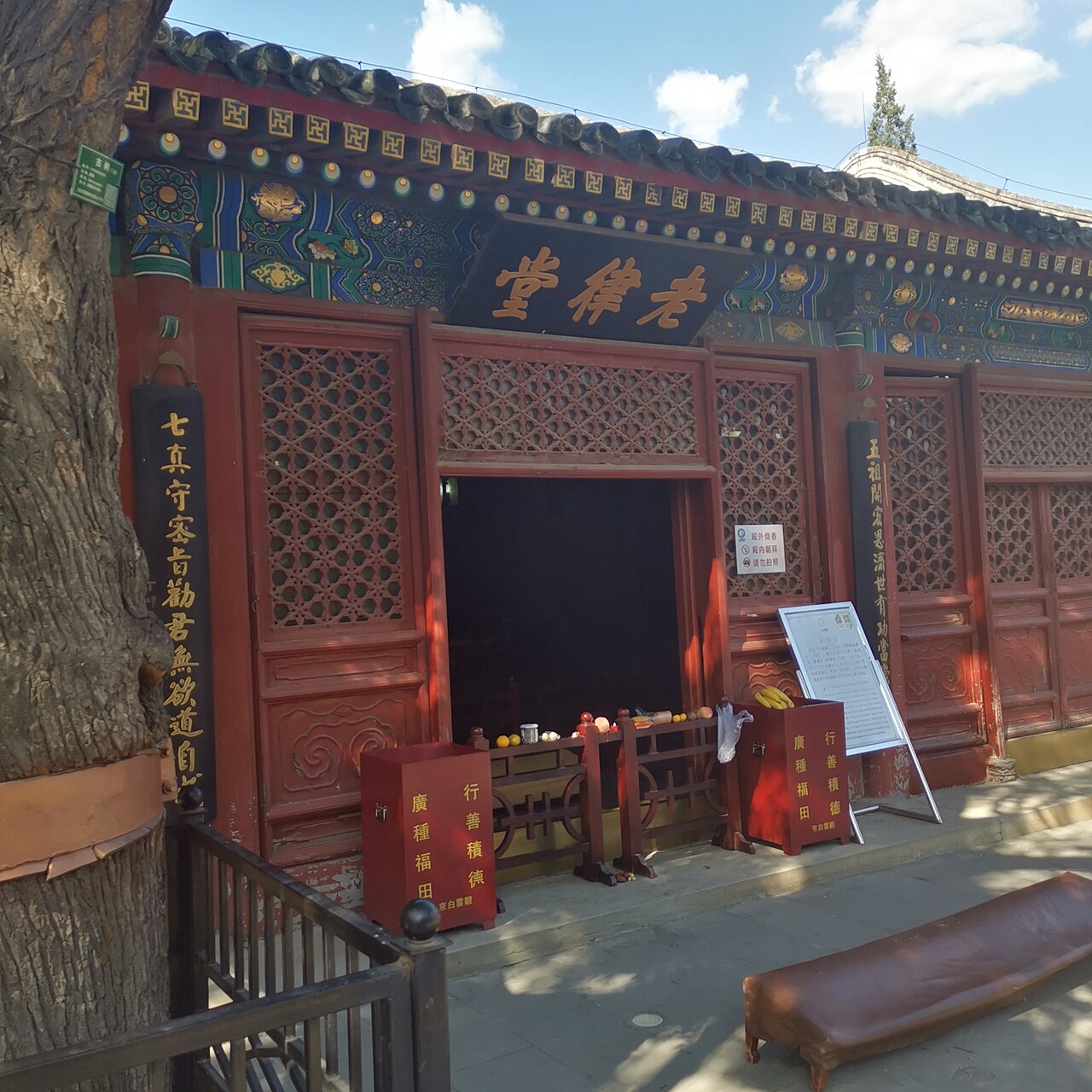 Red latticed facade of Laolü Hall at the White Cloud Temple, its black plaque in gold script above fruit offerings and donation boxes