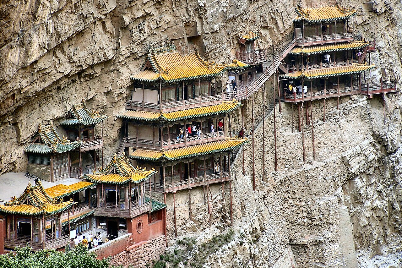 Visitors moving in single file along the galleries of the Hanging Temple, its pavilions standing on slender stilts against the cliff of Jinlong Gorge