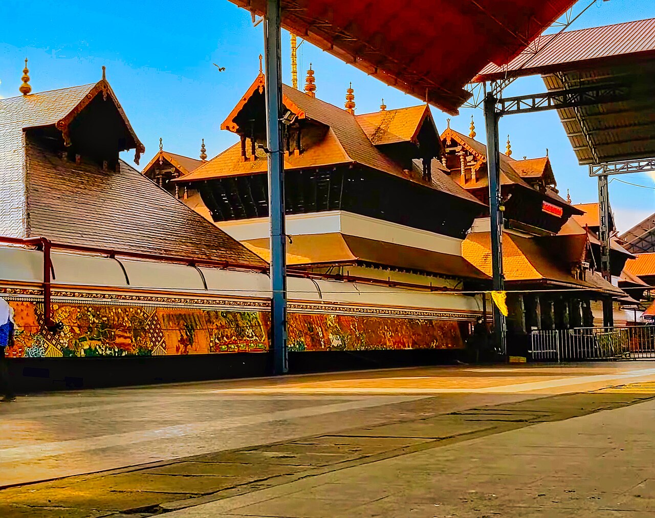 Mural panels running beneath the layered copper roofs along the outer wall of the Guruvayur Temple