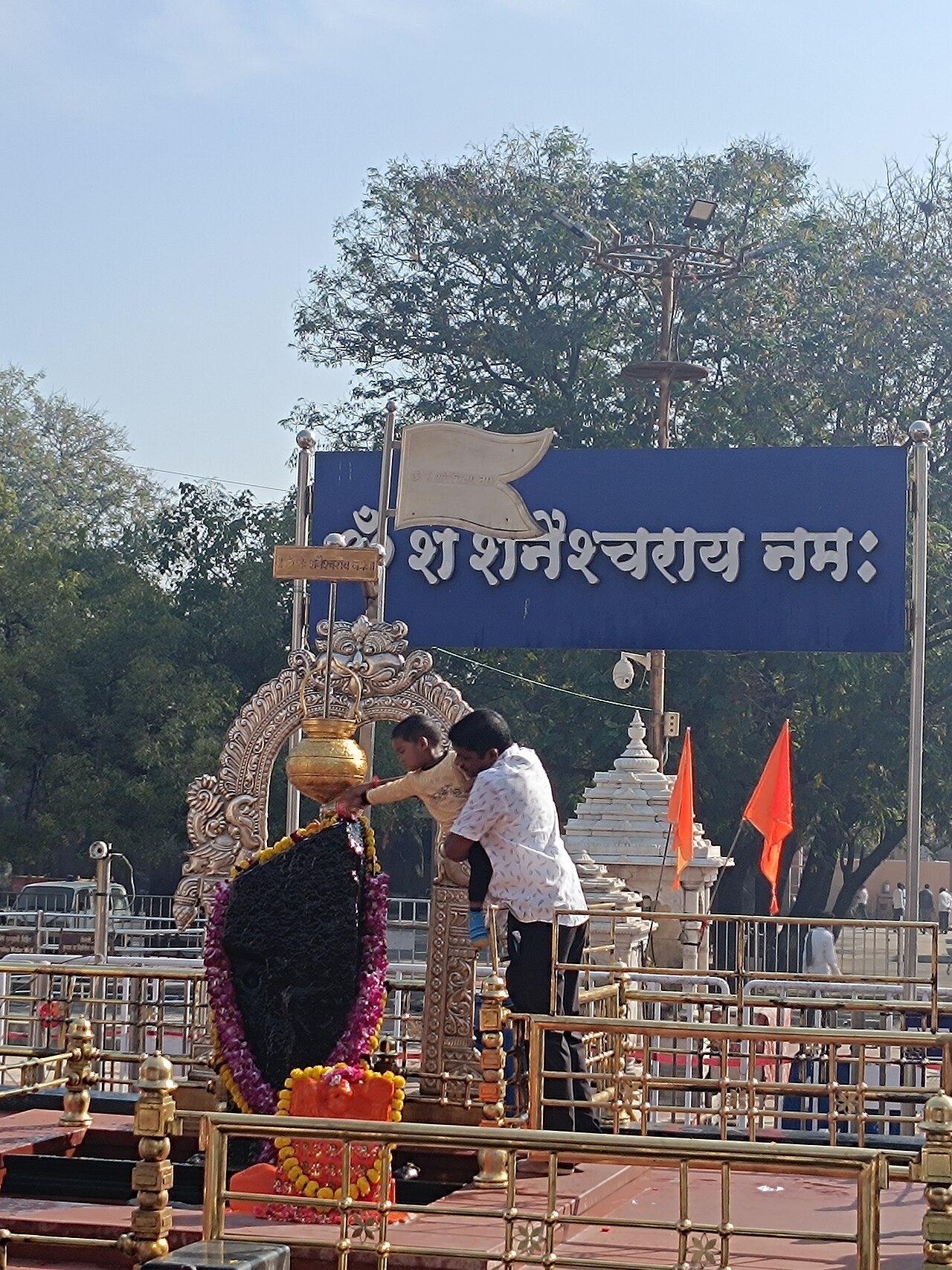 A devotee lifts a child to touch the black slab of Lord Shanidev on the open chauthara at Shani Shingnapur, beneath the golden oil vessel
