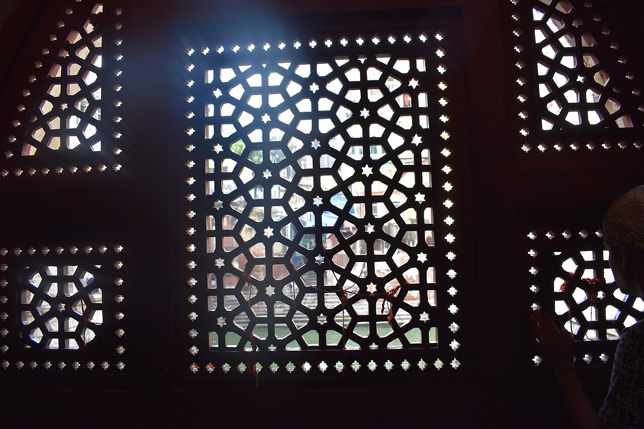 Prayer threads tied into a carved marble jali screen at the dargah, the baoli water visible through the lattice