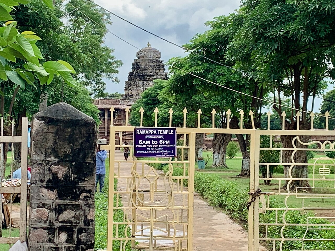 The garden approach to Ramappa Temple, its stepped shikhara visible past the entrance gate and ASI visiting-hours sign