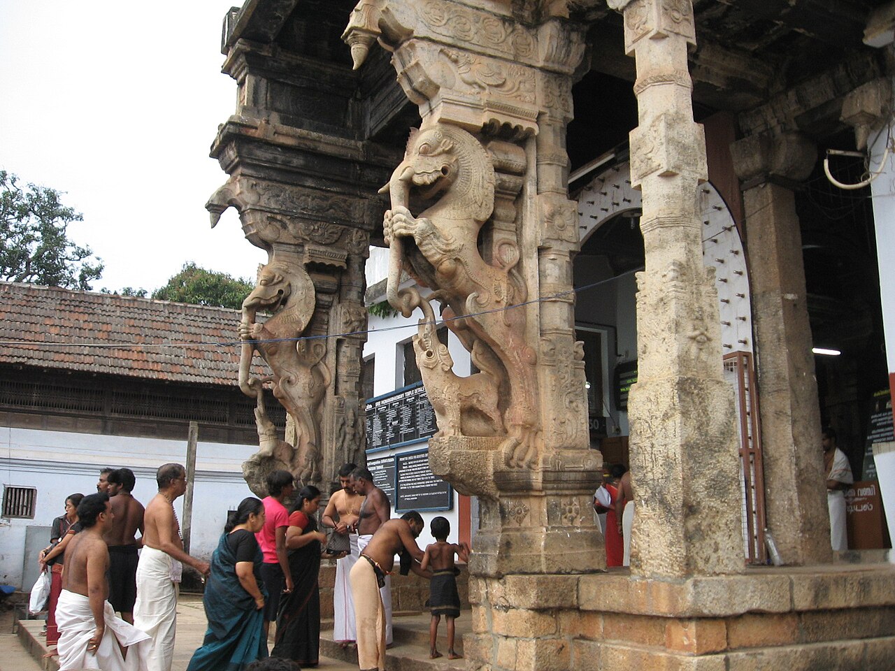 Devotees in mundu and saree passing the carved yali pillars at the entrance of Sree Padmanabhaswamy Temple